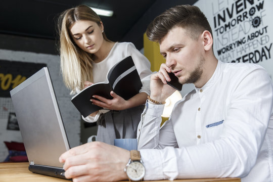 A picture of businessman sitting at table and talking on the phone. He is looking at laptop nad holding hand near it. Girl is standing besides man and writing down. She is concentrated on that.