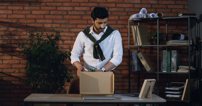 Handsome stylish man office worker packing a box with office stuff from his table and leaving an office with this box in hands as been fired or quitting his job.