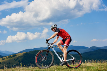 Naklejka na ściany i meble Athlete sportsman bicyclist in professional sportswear and helmet riding cross country bicycle on summer day. Mountains view and cloudy sky on background. Active lifestyle and outdoor sport concept