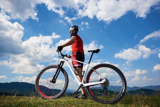Back view of athletic professional rider in sportswear and helmet standing with cross country bicycle, against blue sky with clouds on summer sunny day. Active lifestyle and outdoor sport concept