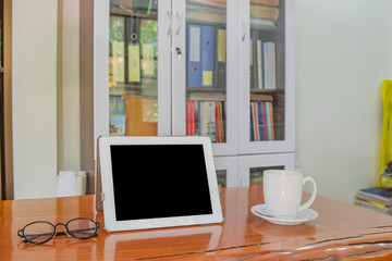 Mock up image of white tablet pc with black blank screen and coffee cup on wooden table in blur office or bookcase background. Ready to work fast and modernize all types of teams in workspace.