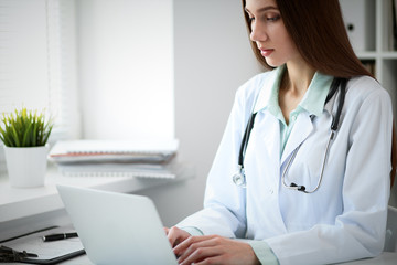 Young female doctor typing on laptop computer while sitting at the table near the window in hospital office