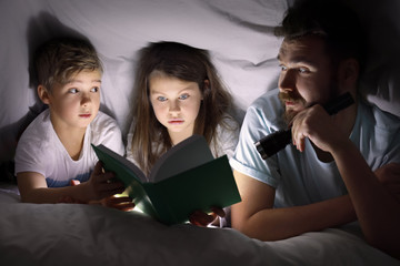 Cute little children with their father reading bedtime story under blanket in evening