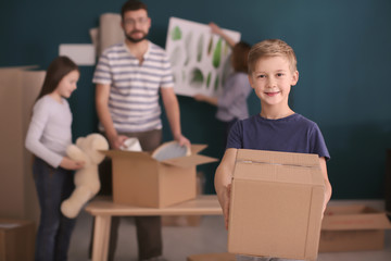 Cute child holding box while his family packing belongings on background. Moving day