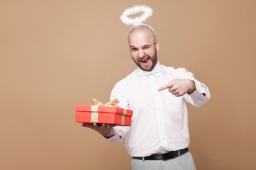 This is for you. Happy funny middle aged bald bearded angel in shirt and white halo on head pointing at red gift and winking with smile. indoor studio shot, isolated on light brown background.