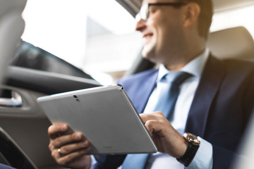 Young businessman using tablet pc while sitting on back seat of a car. Caucasian male business executive by a taxi and looking at digital tablet.
