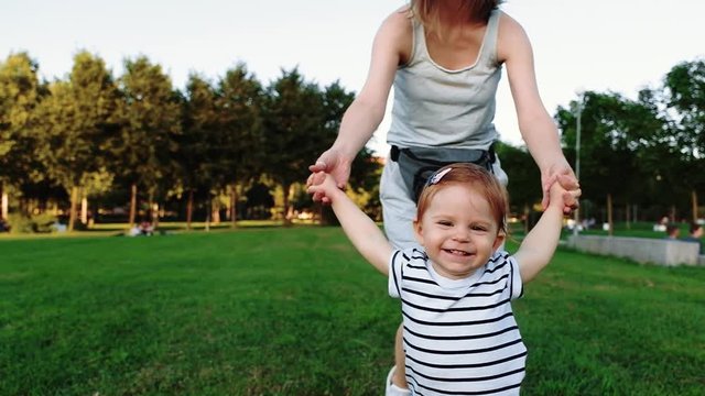 Young pretty mother with her Cute Little laughing baby daughter walking by grass at sunset in a park in suumer day. Slow Motion. Happy childhood. Love and Family