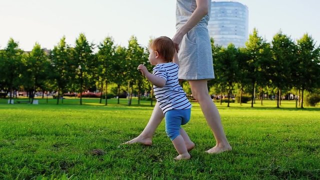 Young pretty mother with her Cute Little laughing baby daughter walking by grass at sunset in a park in suumer day. Slow Motion. Happy childhood. Love and Family