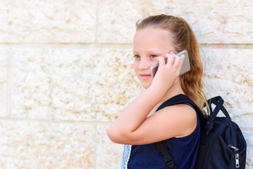Outdoor portrait of happy girl 8-9 year old talking on phone. Cute schoolgirl with backpack talking on cell phone and smiling.