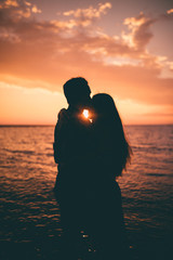 Giovane coppia di ragazzo e ragazza si baciano abbracciandosi di fronte al tramonto rosso fuoco in riva al mare sulla spiaggia. Vista controluce, silhouette. 