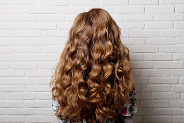 Hairstyle long curls on the head of a brown-haired woman at the back close-up against a white brick wall.