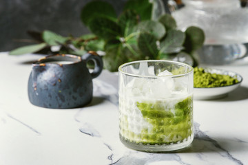 Matcha green tea iced latte or cocktail in glass with ice cubes, matcha powder and jug of milk on...