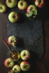 Ripe organic gardening green red apples with leaves and jar of honey on black slate board over dark texture background. Flat lay, space. Autumn harvest.