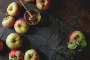 Ripe organic gardening green red apples with leaves and jar of honey on black slate board over dark texture background. Flat lay, space. Autumn harvest.