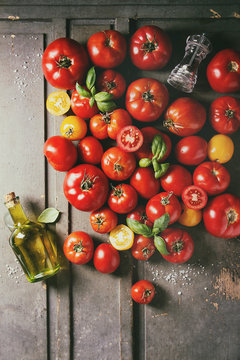 Variety of ripe fresh organic gardening tomatoes different kind and colors with water spot, basil leaves, bottle of olive oil, black pepper and salt over old grey wooden background. Flat lay, space