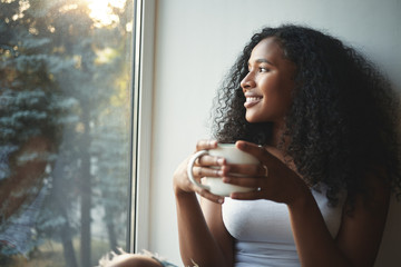 Morning routine. Portrait of happy charming young mixed race female with wavy hair enjoying summer view through window, drinking good coffee, sitting on windowsill and smiling. Beautiful daydreamer