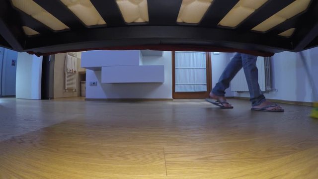 Man wearing flip flops, washes the parquet floor under the bed with a yellow mop