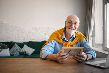 Horizontal shot of cheerful sixty year old stylish businessman wearing rectangular eyeglasses sitting in front of open portable computer, holding family portrait in photo frame and smiling happily