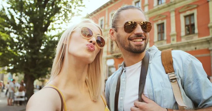 POV of the young cheerful Caucasian couple posing and smiling while taking selfie photos while standing on a town street. Close up.