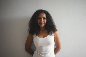 Fashionable young dark skinned woman wearing white tank top smiling and keeping hands behind her...