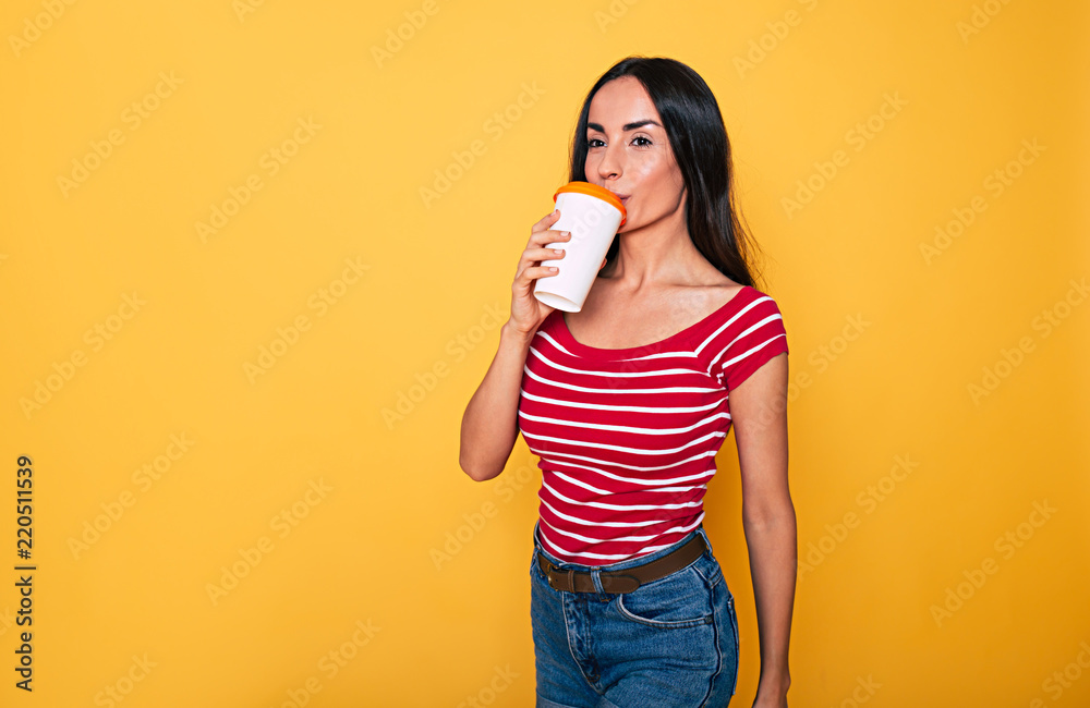 Wall mural smiling attractive woman in casual clothes with coffee cup in hands posing over yellow background