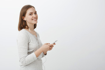 Profile shot of entertained good-looking carefree adult female with short brown haircut holding smartphone gazing aside with satisfied cute smile listening music in earphones over gray background