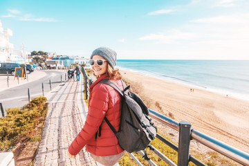 Fototapeta na wymiar Beautiful young woman in hat and sunglasses walking in winter on the winter coast of the Atlantic Ocean in the Algarve, Portugal