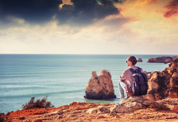 Beautiful young woman traveler sits on the rock and looks at the sunset over the ocean in the...