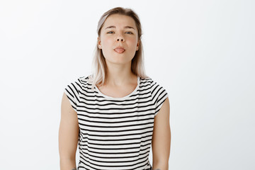 Indoor shot of playful good-looking woman in striped t-shirt, tilting head and showing tongue, being childish and in great mood, playing with friend and making faces while being bored during classes