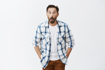 Guy feeling surprised finding money in pocket of old trousers. Portrait of amazed and thrilled good-looking man with beard, folding lips while say wow, gazing excited at camera, posing over grey wall