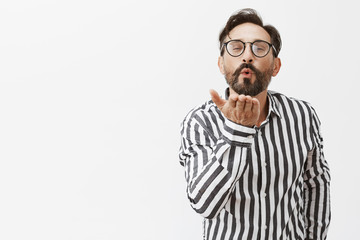 Obraz premium Mwah my charming love. Romantic good-looking adult man with beard, sending kiss by air, folding lips and holding palm near mouth, standing over gray background and showing affection to wife