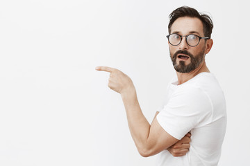 Wow, it is masterpiece. Portrait of charmed and impressed good-looking mature male in glasses and t-shirt, standing in profile, turning to camera to express amazement, pointing left with index finger