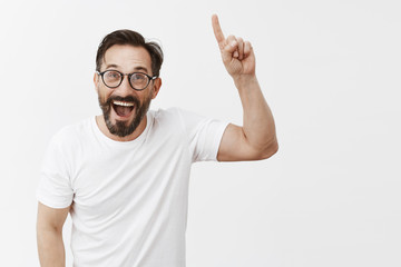 Excited happy handsome man with beard in trendy eyewear and t-shirt, raising index finger to point up in sky or show eureka gesture, smiling joyfully, rejoicing and triumphing, being right