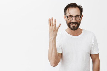 I have four kids. Portrait of good-looking charming happy male model with beard and moustache in black glasses and t-shirt, showing fourth number and smiling broadly at camera over grey wall