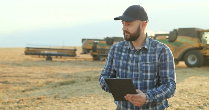 Caucasian man in a hat standing in the field and calculating amount of grain while noting in the documents while machines gathering harvest behind him.