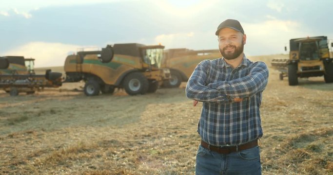 Portrait shot of the agricultural businessman standing in the field and posing to the camera. Gathering of the harvest on the background.