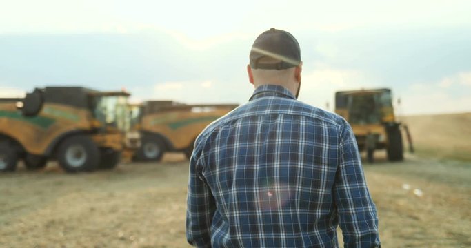 Back view on the man in the plaid blue shirt and hat walking in the field to the combines machinery. Rear.