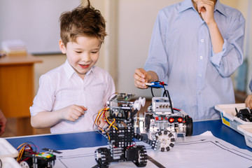 Fair haired cheerful little boy making a robot from metal parts and microcircuits, his brother and...