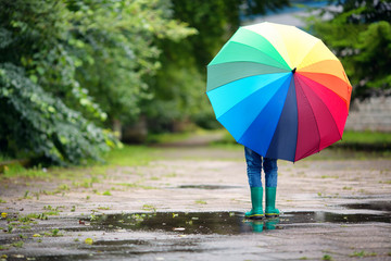 Child walking in wellies in puddle on rainy weather. Boy holding colourful umbrella under rain in summer © candy1812