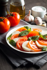 Photo of Caprese Salad with tomatoes, basil, mozzarella, olives and olive oil on wooden background. Italian traditional caprese salad ingredients. Mediterranean, organic and natural food concept.