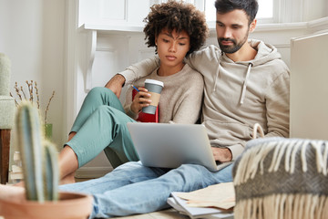 People, leisure, mutliethnic relationship concept. Lovely mixed race couple spend free time at home, dressed casually, embrace, sit in front of laptop computer, watch film and drink takeaway coffee