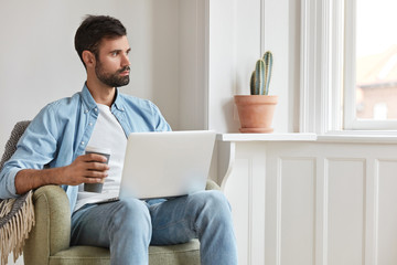 Pensive bearded blogger, sits in armchair in cozy room, works on laptop computer, drinks takeaway...