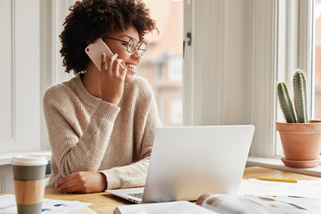 Horizontal shot of adorable cheerful dark skinned young lady calls to customer service, checks...