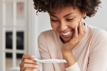 Close up shot of happy future mother smiles broadly when finds out positive results of pregnancy test, cant believe in such good news, poses against cozy domestic background. Yes, I am pregnant!