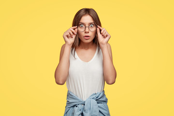 Emotive horrified young female model stares through spectacles, dressed in casual outfit, cant believe her eyes, keeps hands on rim, poses over yellow wall, terrified with accident or awful news