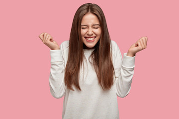Successful brunette girl closes eyes from happiness, keeps raised fists, dressed casually,...