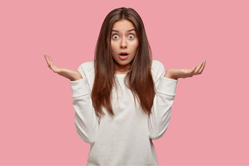 Shocked brunette young woman looks with puzzlement at camera, feels surprised and uncertain, stares at camera and shruggs shoulders, wears casual loose white sweatshirt, stands against pink wall.