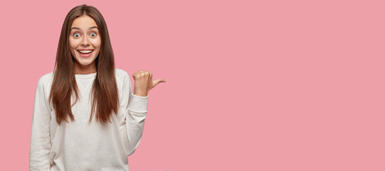 Horizontal portrait of amazed beautiful happy lady dressed in casual clothes, points with thumb at free space, suggests you visit this place, isolated over pink background. Advertisement concept
