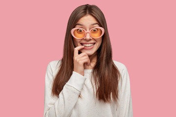 People and style concept. Positive emotive smiling schoolgirl, wears stylish sunglasses, keeps fore finger near lips, listens something with great interest and intrigue, stands over pink studio wall