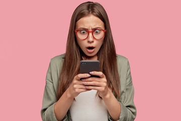 Shocked woman with terrific expression, stares at mobile phone, being stunned with sudden break of cellular, dressed in fashionable casual clothing, stands indoor. People, reaction, technology concept